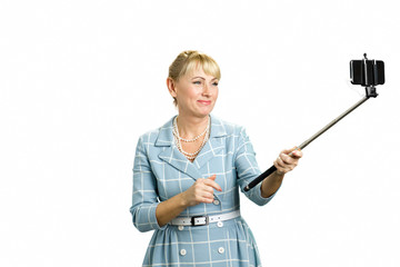 Beautiful mature lady taking self portrait. Attractive middle aged woman taking selfie with selfie stick standing on white background.