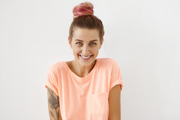 Charming playful coquettish young European female with pink hair bun smiling shyly at camera as if...