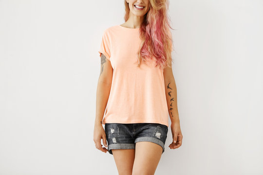 Cropped studio portrait of smiling joyful young woman with long pinkish hair and stylish tattoos on arms posing at white blank wall wearing ripped jeans shorts and oversize peach color t-shirt