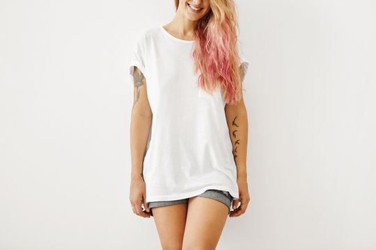 Unrecognizable beautiful young Caucasian woman with tattooed arms smiling broadly dressed in blank white t-shirt. Picture of stylish trendy looking teenage girl posing in studio in trendy summer look