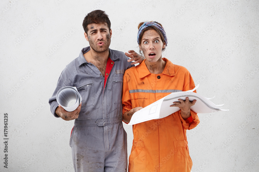 Wall mural displeased female and male construction workers, hold blueprint, have untidy faces, look with discon