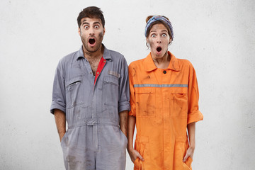 Repair and renovation concept. Professional male and female technicians look with bugged eyes and opened mouth, realize their fault at work. Astonished young repairers in overalls with dirty faces