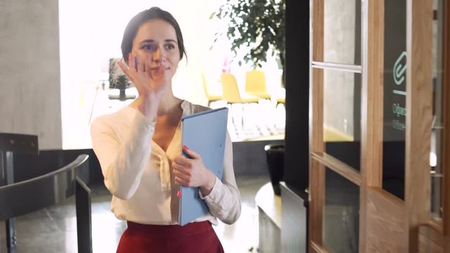 young working woman dressed in white blouse and short skirt go with document case in hands through the office greeting with collegues during sunny day