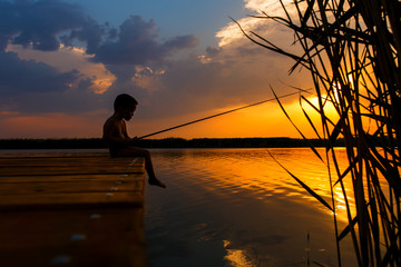 Little boy siting on wooden dock and fishing at sunset.