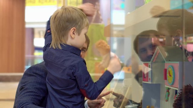 The boy shows on a toy which he wants for Christmas. The little fair-haired boy looks at the range of toys in toy store. The boy has leaned the elbows on glass of shop in Christmas vanity. Child On