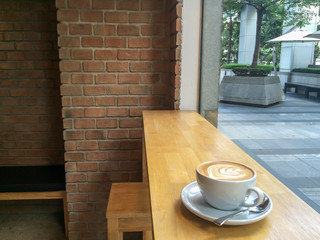 Cup of morning coffee on wooden table in modern coffee shop / cafe restaurant with brick wall with outdoor view in morning light, with copy space in background.