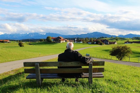 Älterer Mann sitzt auf einer Bank und genießt den Panoramablick auf die Berge, Allgäu, Bayern