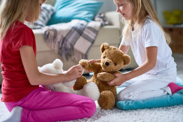 Side view of pretty little sisters playing with plush teddy bears while sitting on cozy carpet at spacious living room