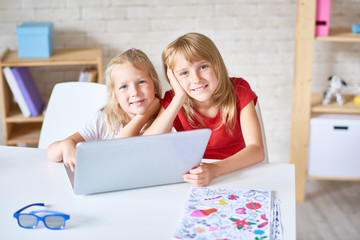 Group portrait of cheerful little sisters posing for photography with toothy smiles while  watching their favorite cartoon on laptop, interior of cozy bedroom on background