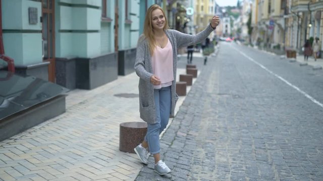 young attractive woman is standing on the road and trying to catch a car hitchhiking. a woman dressed in blue jeans and a gray knitted sweater. the girl rests while walking around the old city. cool