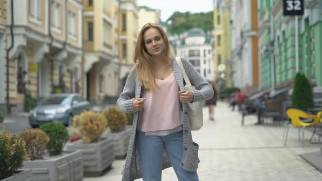 young attractive woman is walking around the city among the buildings. laughs cheerfully. a woman dressed in blue jeans and a gray knitted sweater. cool autumn weather 4 k