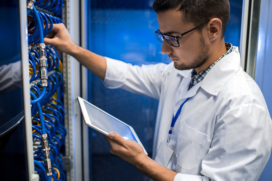 Portrait of young man wearing lab coat working with supercomputer connecting blade server cables and  checking data on digital tablet