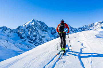 Mountain ski walking up along a snowy ridge with skis in the backpack. In background blue cloudy sky and shiny sun and Tre Cime, Drei Zinnen in South Tirol, Italy. Adventure winter extreme sport.