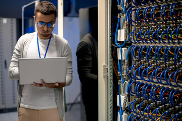 Portrait of young man with laptop standing by server cabinet while working with supercomputer in...