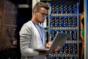 Side view  portrait of young man with laptop standing by server cabinet while working with...