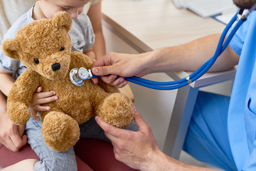 Unrecognizable pediatrician examining plush teddy bear with help of stethoscope while his little patient having check-up