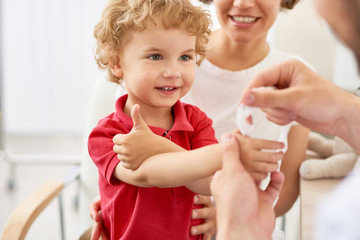 Waist-up portrait of smiling little boy showing thumb up while pediatrician putting bandage on...
