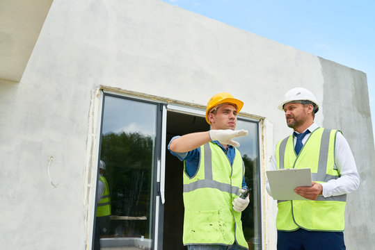 Handsome young foreman wearing hardhat and reflective jacket discussing results of accomplished work with bearded middle-aged investor, unfinished building on background
