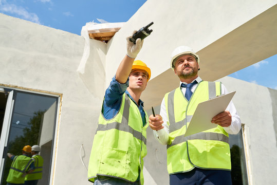 Low angle view of confident bearded businessman wearing suit and hardhat talking to young foreman while checking construction phase of building