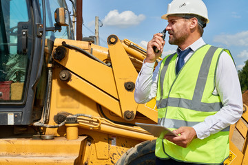 Profile view of handsome middle-aged construction worker wearing suit and hardhat talking via walkie-talkie while standing at road roller, he holding digital tablet in hand