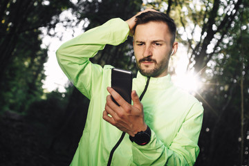 Bearded pensive athlete with bluetooth headphones looks at smartphone screen with workout sport statistics