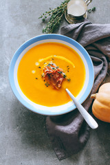A plate of pumpkin soup with cream, prosciutto and thyme, on a gray background, top view