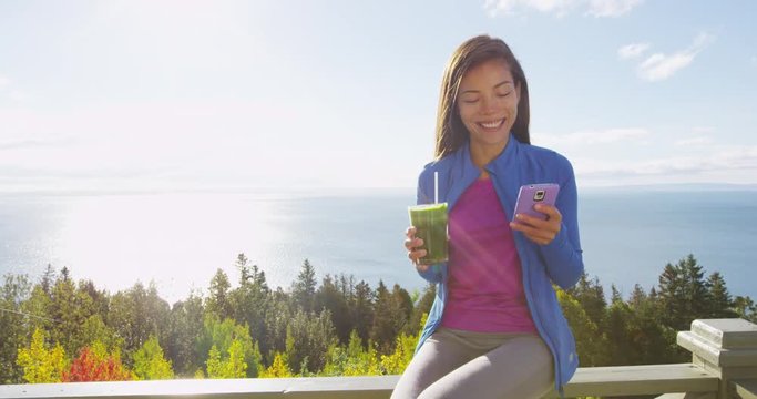 Healthy eating girl drinking green smoothie detox using cell phone in autumn fall foliage nature retreat. Happy woman using smartphone app on weight loss diet vegan nutrition cleanse.