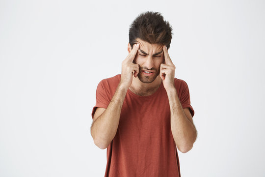 Portrait of attractive young spanish male wearing red t-shirt with stressful and frowny face, squeezing head with hands having headache after sleeping only for few hours.