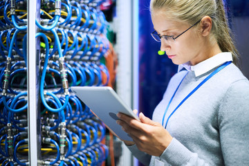 Portrait of young woman wearing glasses using digital tablet standing against server cabinets in...