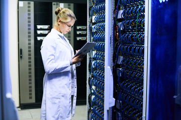 Side view portrait of young woman wearing lab coat  working with supercomputer inputting data to digital tablet standing by server cluster
