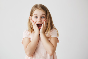 Small blonde girl with blue eyes lookin at camera, holding face with hands and opened mouth expressing excitement. Kid getting happy after receiving birthday present from mother.