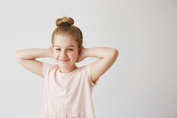 Beautiful young miss with blue eyes and light hair in pink t-shirt posing with cute bun hairstyle and hands behind head for family photo.