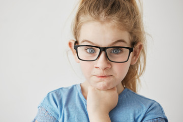 Fototapeta premium Close up of little cute girl with big blue eyes and light hair looking at camera with angry face expression and raised eyebrows in glasses.