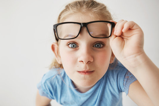 Close up portrait of curious little girl with big blue eyes standing close and looking in camera, holding glasses with hand.