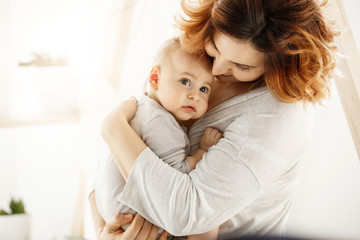Cute newborn kid frightenedly looks aside while good-looking young mother gently huggs baby...