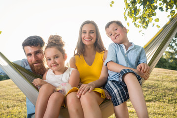 Family portrait with a beautiful mother of two playful children swinging in a hammock while looking at camera next to her husband outdoors in summer