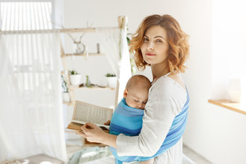 Portrait of a beautiful young mother with sleeping son on chest and book in hands. Woman turns head...