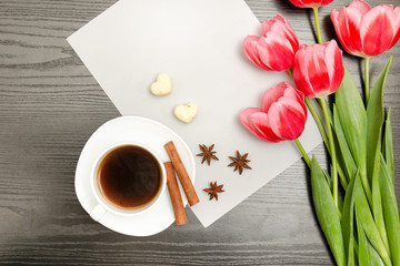 Pink tulips on a blank sheet of paper, mug of coffee and spices, black wooden background. top view