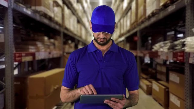 Caucasian warehouse worker working with a tablet computer in the warehouse corridor and wearing blue uniform