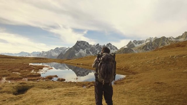 man photographer hiker shoot photo near lake to snowy mounts.Behind view. Sunny autumn day Hiking in colorful red forest wild nature mountain outdoors.Alps Devero lake park. 4k video