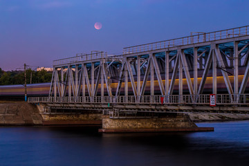 Fototapeta na wymiar Train is passing through railway bridge, long exposure. Moon eclipse on the sky 