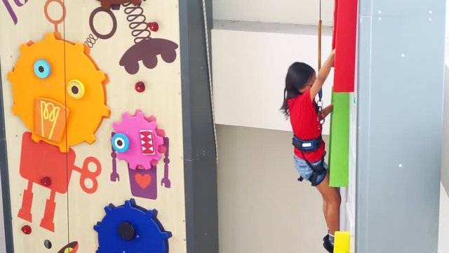 Video footage of a little girl climbing a rock wall indoors while wearing a safety rope
