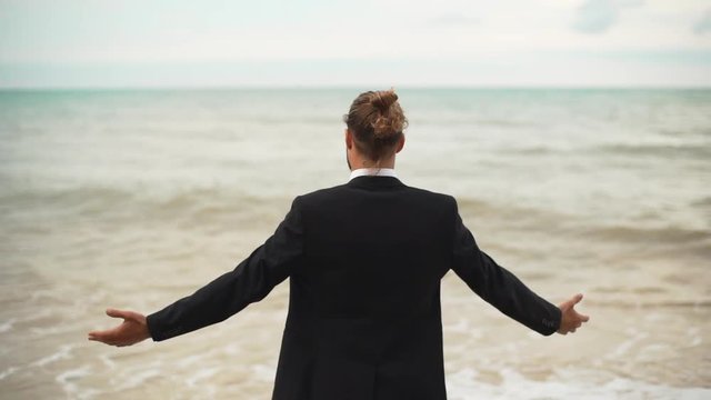 businessman with tied hair greeting sea with open arms backside to camera slowmotion