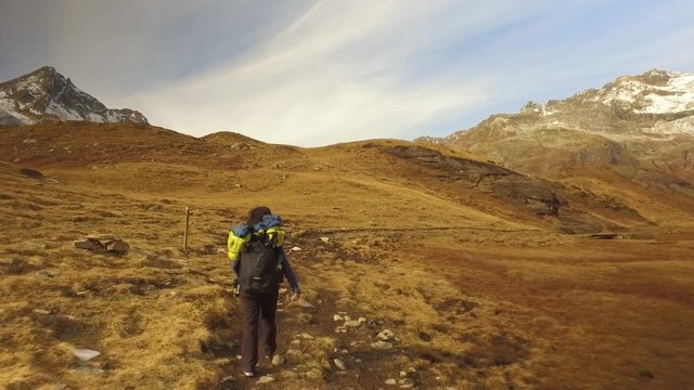 woman hiker walking on fall fields path to snowy mounts.Following behind view. Sunny autumn day Hiking in colorful red forest wild nature mountain outdoors.Alps Devero lake park. 4k video