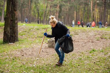 Young blonde girl picking up the garbage and putting it in a black garbage bag on a natural background. Ecology protection concept/Woman volunteer collecting garbage in park - Powered by Adobe