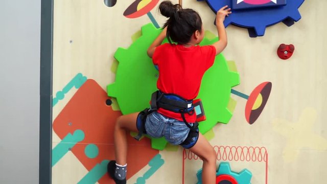 Video footage of a little girl climbing a fun wall indoors to train her courage, strength, endurance and agility while wearing safety rope
