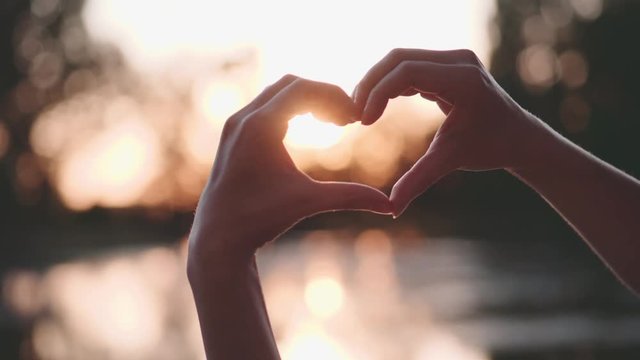 Woman Shapes Heart with Hands over Sun on Sunrise or Sunset. Slow Motion 120 fps, 4K.  Female hands making a heart shaped sign against the sun, near the lake, outdoors. Hope, Happiness, Love Concept. 