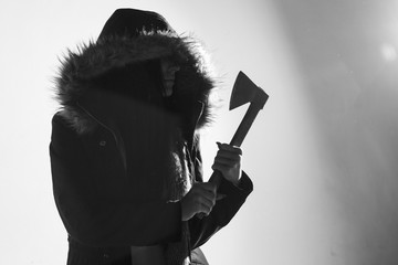Spooky woman in a coat with fur hood, standing holding an axe. Over a white wall. Converted to black and white, grain added.