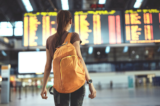 Young woman with small backpack as a hand luggage in international airport looking at the flight information board, checking her flight