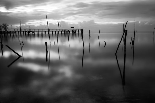 Surreal long exposure view of a lake, with wooden poles on perfectly still water and a pier in the background